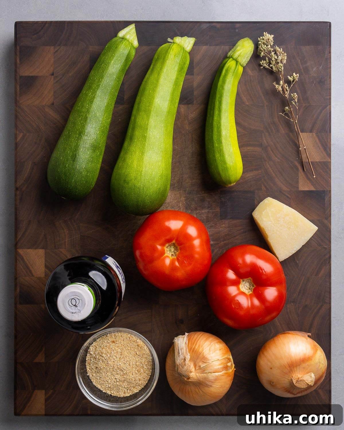 Zucchini Parmesan Bake 3 Ingredients for Italian zucchini casserole: zucchini, Sicilian oregano, olive oil, tomatoes, Pecorino Romano cheese, breadcrumbs, and onions on a wooden board.