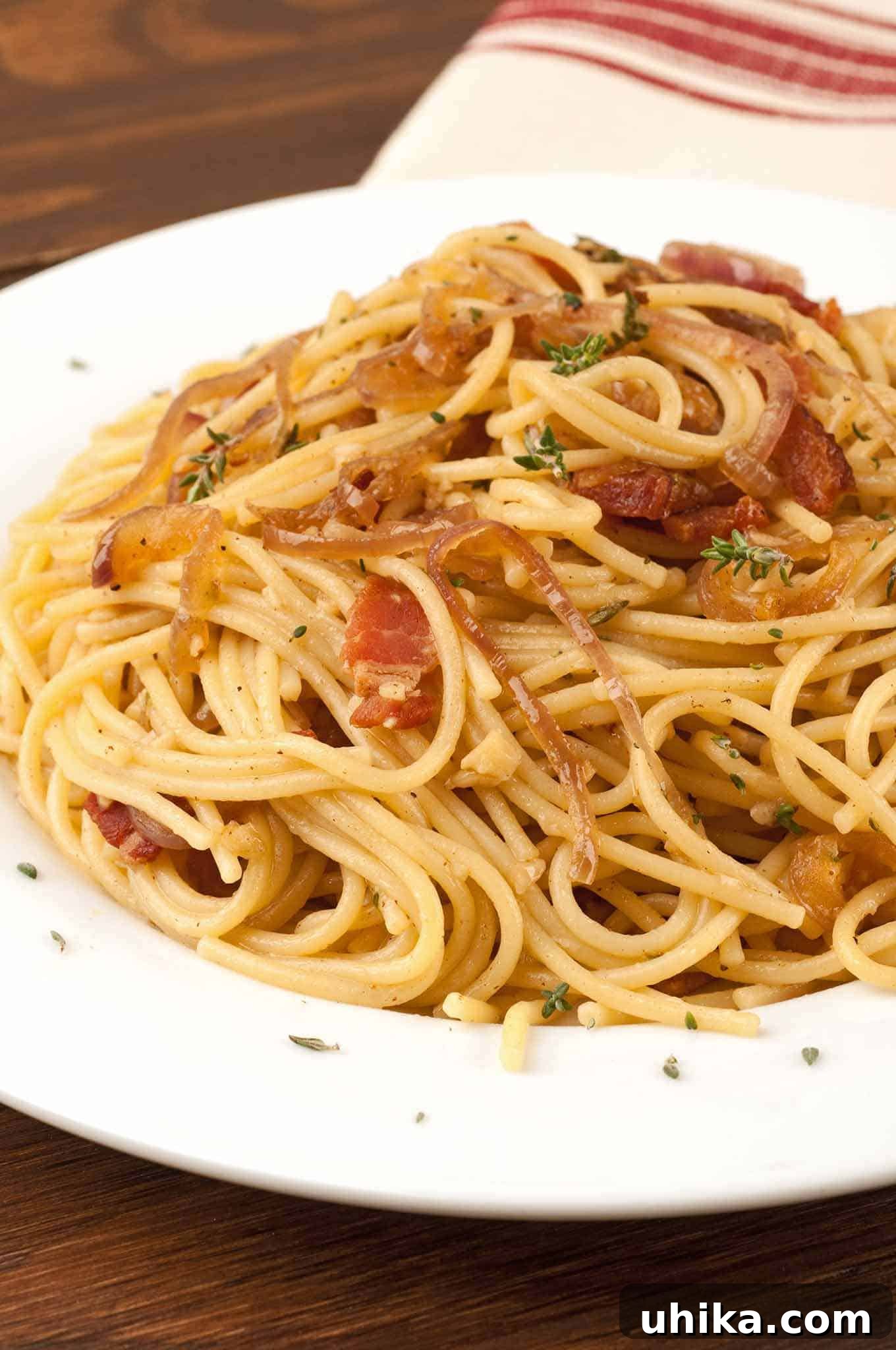 Aromatic Thyme, Bacon, and Onion Spaghetti 2 Spaghetti tossed with crispy bacon, caramelized onions, and fresh thyme on a white dish, a striped towel in the background.