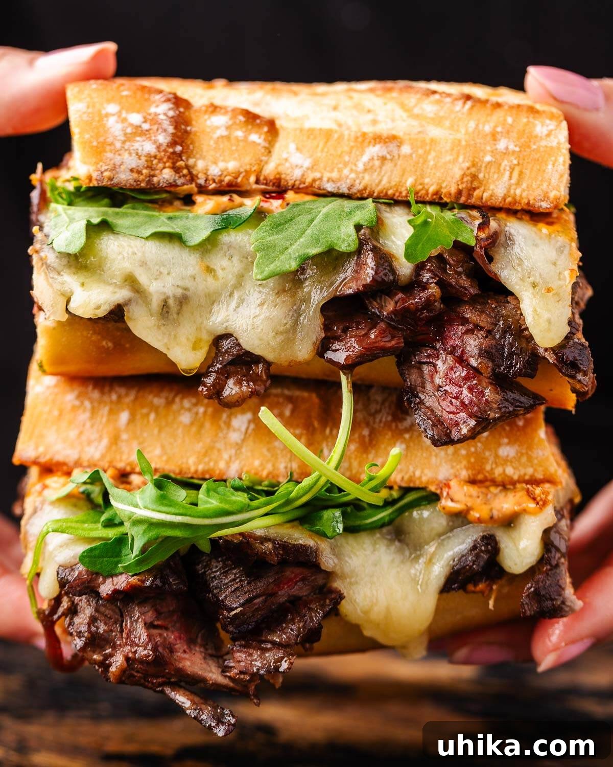 The Ultimate Steak Sandwich 2 Two halves of a delicious steak sandwich, showcasing tender steak, melted cheese, and fresh greens, held in hands.
