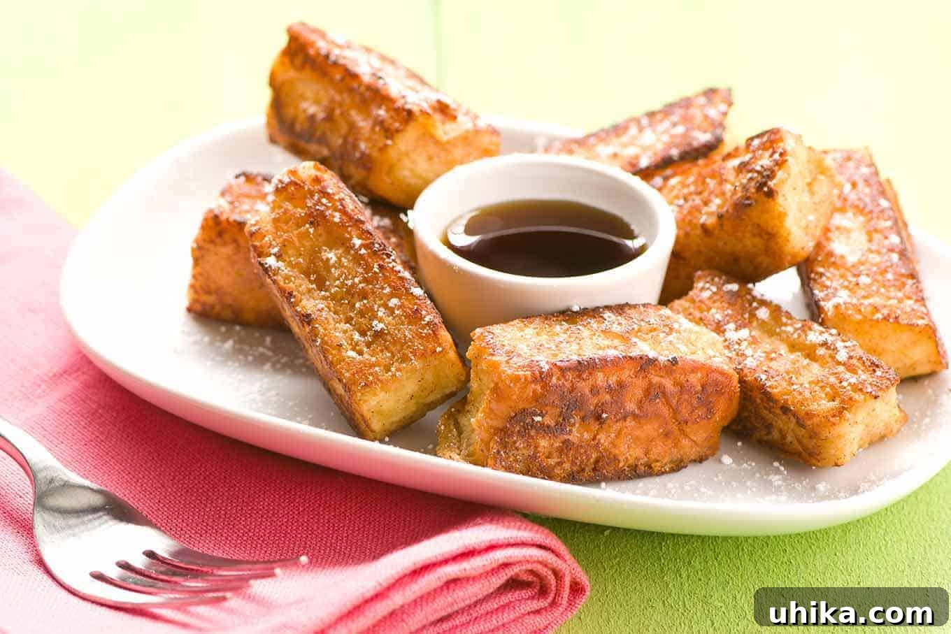 A beautiful serving of French toast sticks made with Hawaiian rolls, elegantly dusted with powdered sugar, and accompanied by a small bowl of golden maple syrup for dipping.