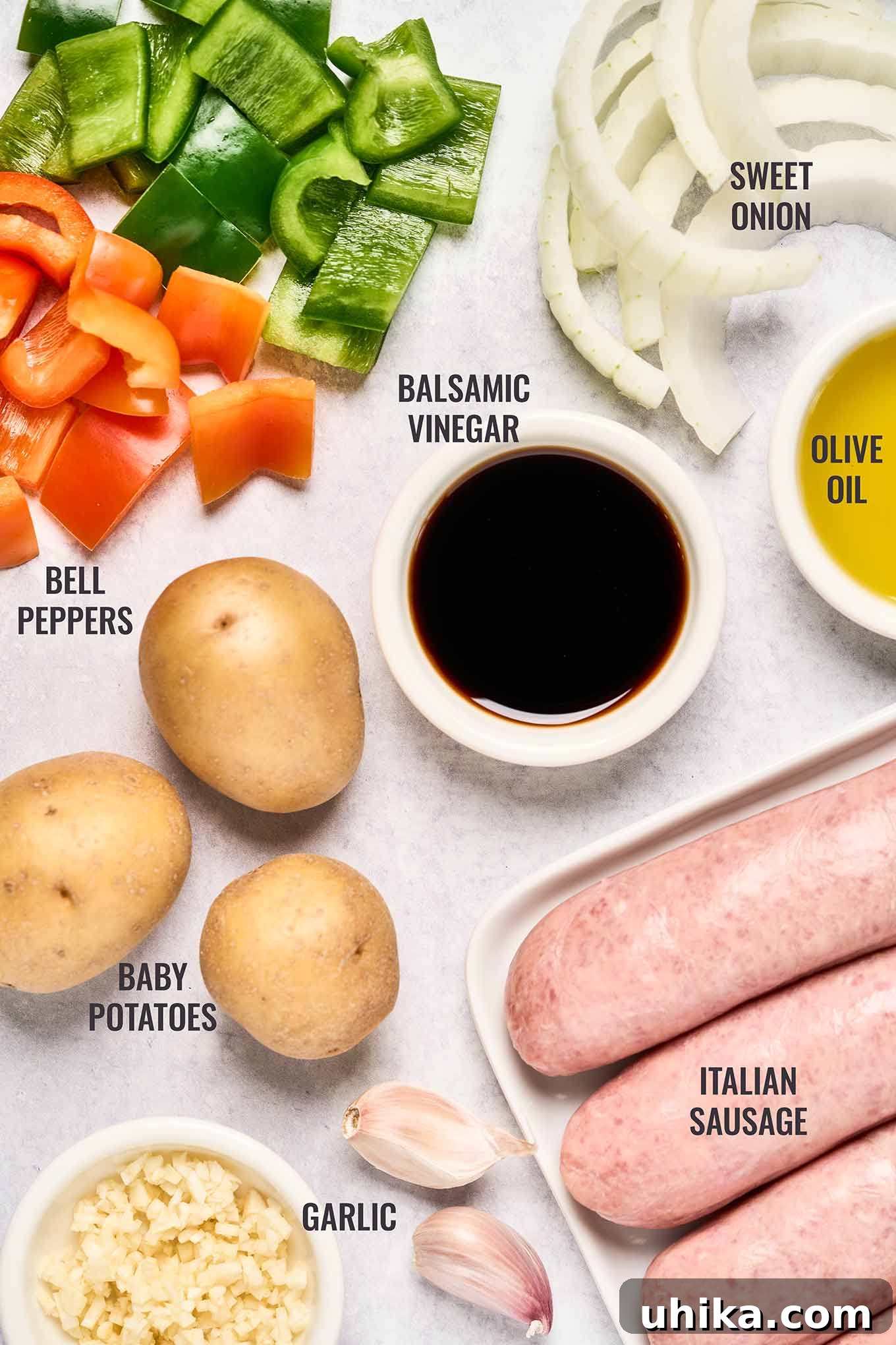 A labeled, overhead view of the ingredients needed for baked Italian sausage dinner; Italian turkey sausage, sliced sweet onion, cubed red and green bell peppers, garlic, balsamic vinegar, and baby yellow potatoes.