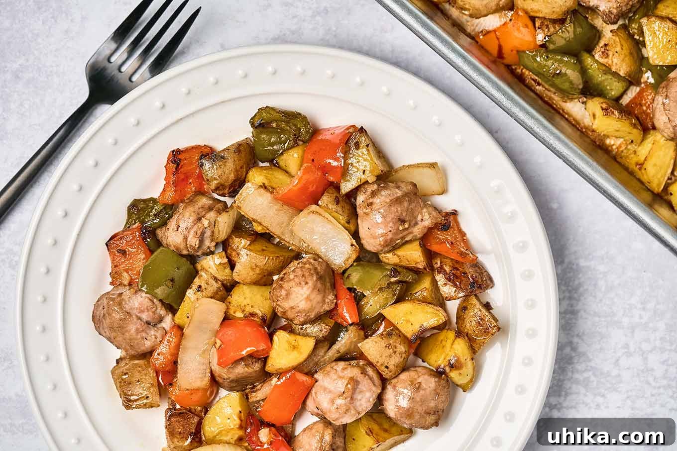 A single serving of baked Italian sausage with bell peppers, potatoes, and onions on a round white plate with the sheet pan it was served from in the upper right, dinner fork on the left.