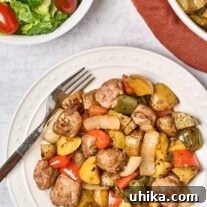A round white dinner plate with with balsamic glazed baked Italian sausage, bell peppers, and onions, a dinner fork to the side and a serving bowl with more of the main dish and a bowl of salad with grape tomatoes in the background.
