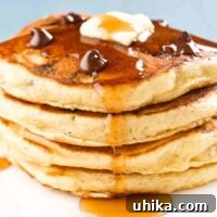 A stack of four chocolate chip pancakes topped with a few extra chocolate chips, a pat of butter, and a drizzle of maple syrup.
