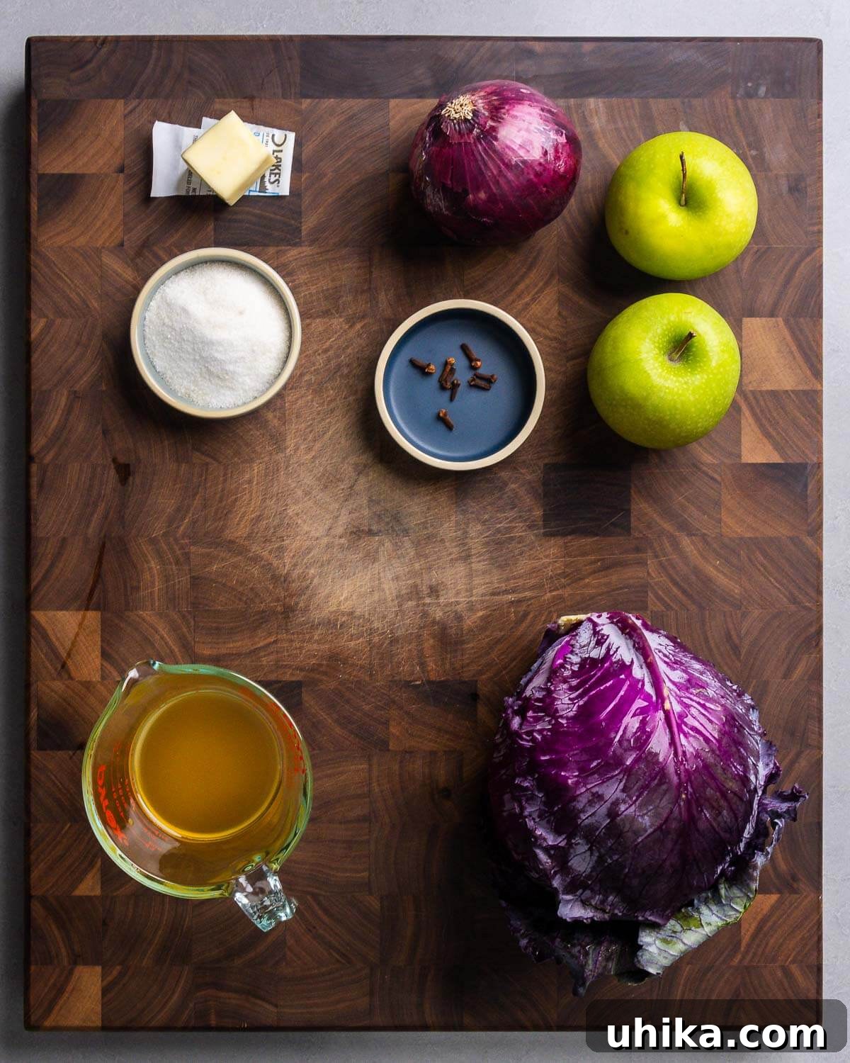 Classic German Red Cabbage 3 Ingredients shown: butter, sliced red onion, Granny Smith apples, sugar, whole cloves, cider vinegar, and a head of red cabbage.