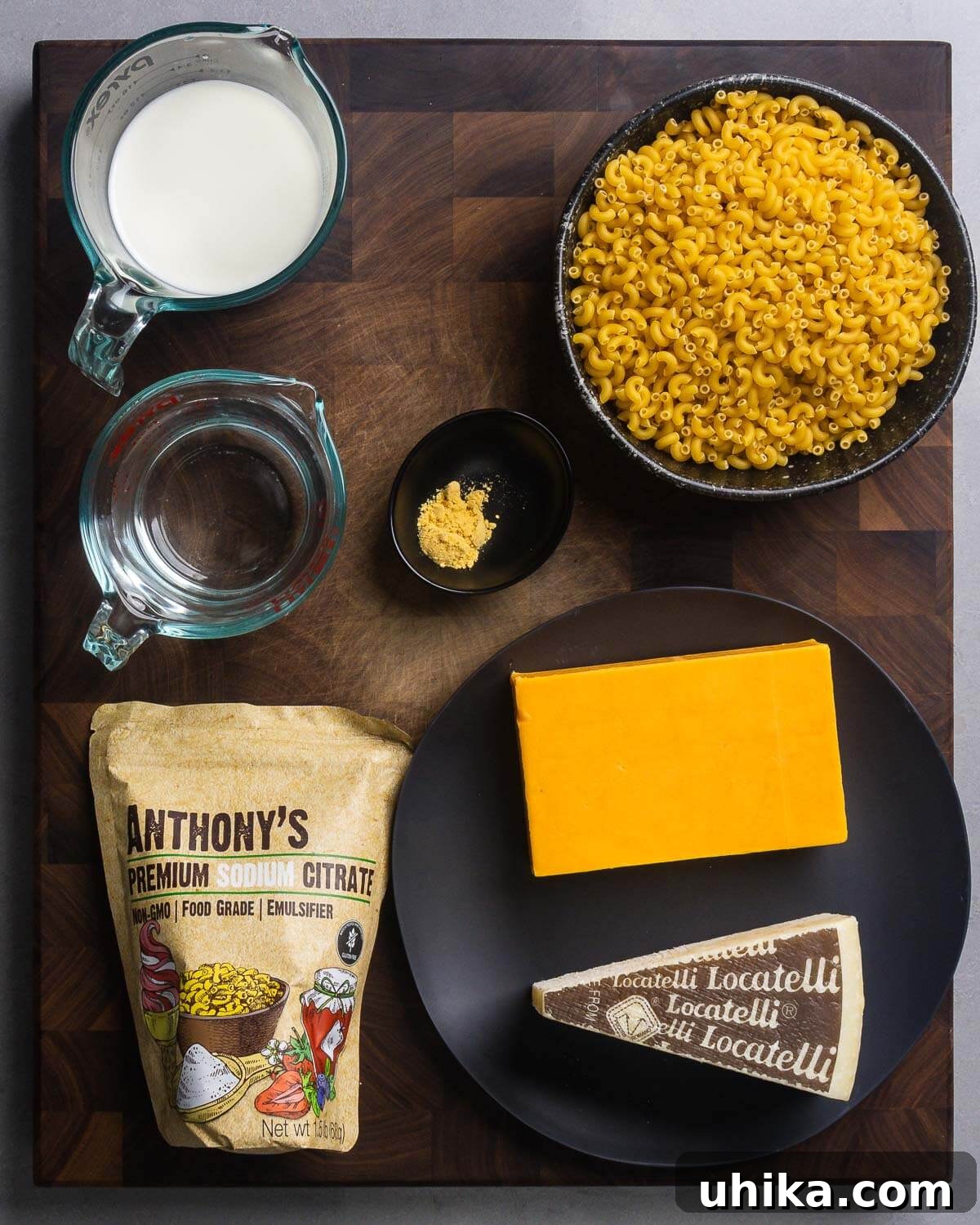 A flat lay photograph of all ingredients laid out: milk, water, dry mustard, uncooked elbow macaroni, a small container of sodium citrate, a block of sharp cheddar cheese, and a block of Pecorino Romano cheese.