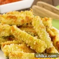 Crispy oven-fried zucchini sticks coated in panko crumbs and grated Parmesan stacked on a serving plate with marinara dipping sauce in the background.