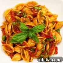 A vibrant serving dish filled with shell pasta, generously tossed with bright red sun-dried tomatoes, fresh green basil leaves, and a glistening balsamic glaze.