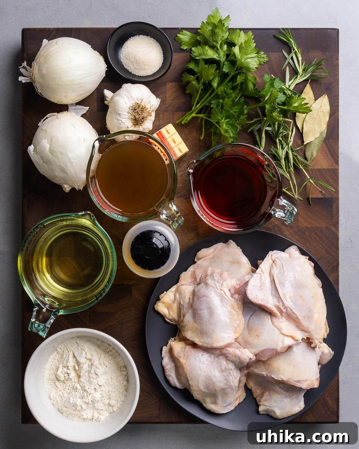 Zesty Italian Vinegar Chicken 3 A flat lay photograph displaying all ingredients for Italian vinegar chicken: onions, granulated sugar, garlic cloves, cold butter, fresh parsley, rosemary sprigs, bay leaves, rich chicken stock, red wine vinegar, dry white wine, olive oil, bone-in chicken thighs, and all-purpose flour.