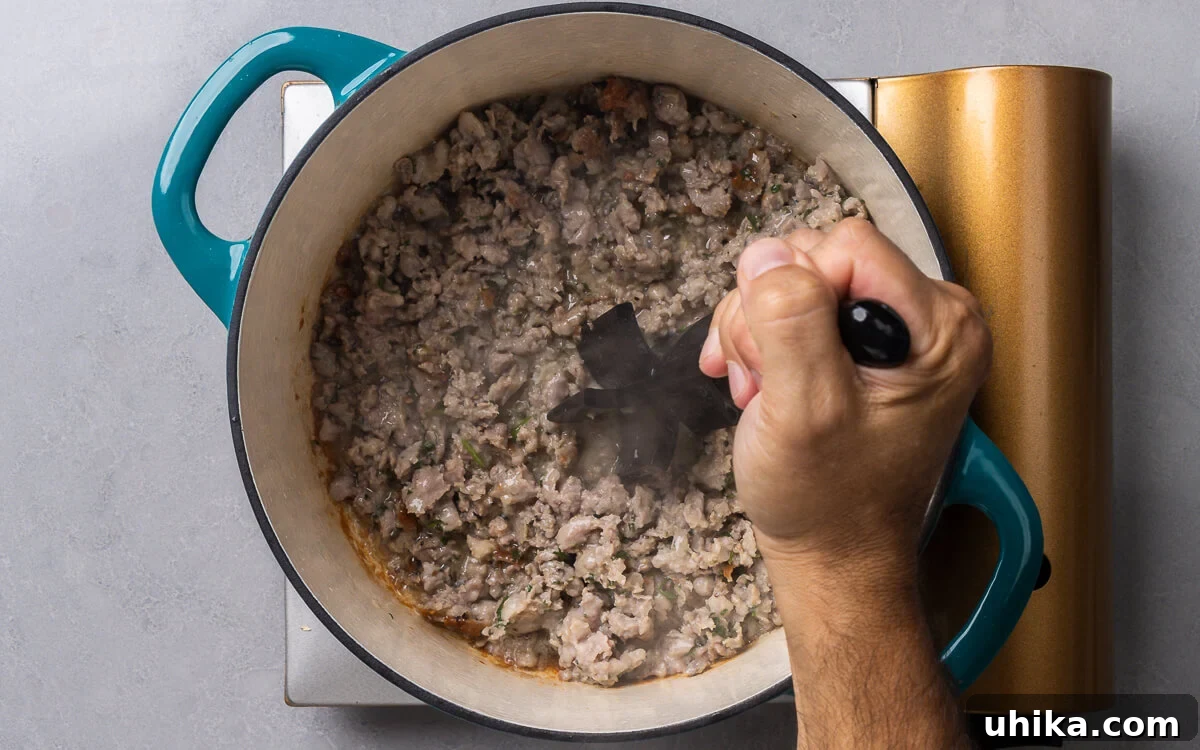 10 Must-Have Kitchen Tools for Your Fall Cooking 5 A hand confidently holding a heat-resistant meat masher over a pot of sizzling sausage, efficiently breaking it apart.