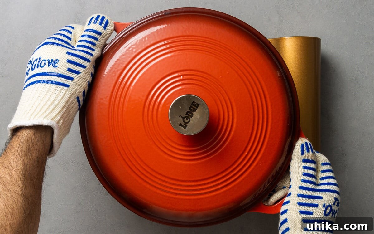 10 Must-Have Kitchen Tools for Your Fall Cooking 6 Two hands, protected by bright orange oven gloves, carefully removing a steaming hot Dutch oven from the oven.