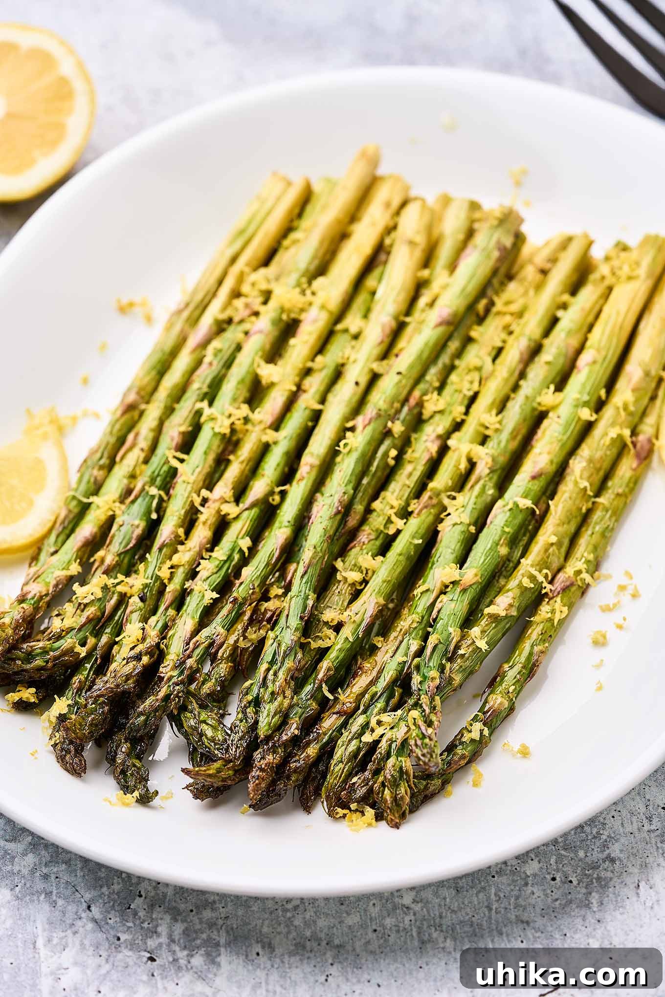 Zesty Lemon Roasted Asparagus 6 Spears of lemon roasted asparagus topped with lemon zest on a white oval serving plate garnished with lemon slices.