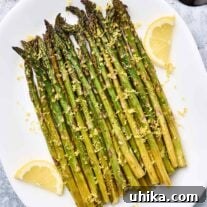A serving plate of lemon roasted asparagus topped with lemon zest and garnished with lemon slices, a black serving fork and half a lemon in the background.