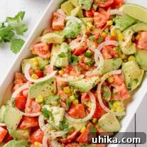 A serving platter of avocado chunks, tomatoes, corn kernels, sliced onion, and cilantro, tossed in creamy lime dressing and garnished with lime wedges.