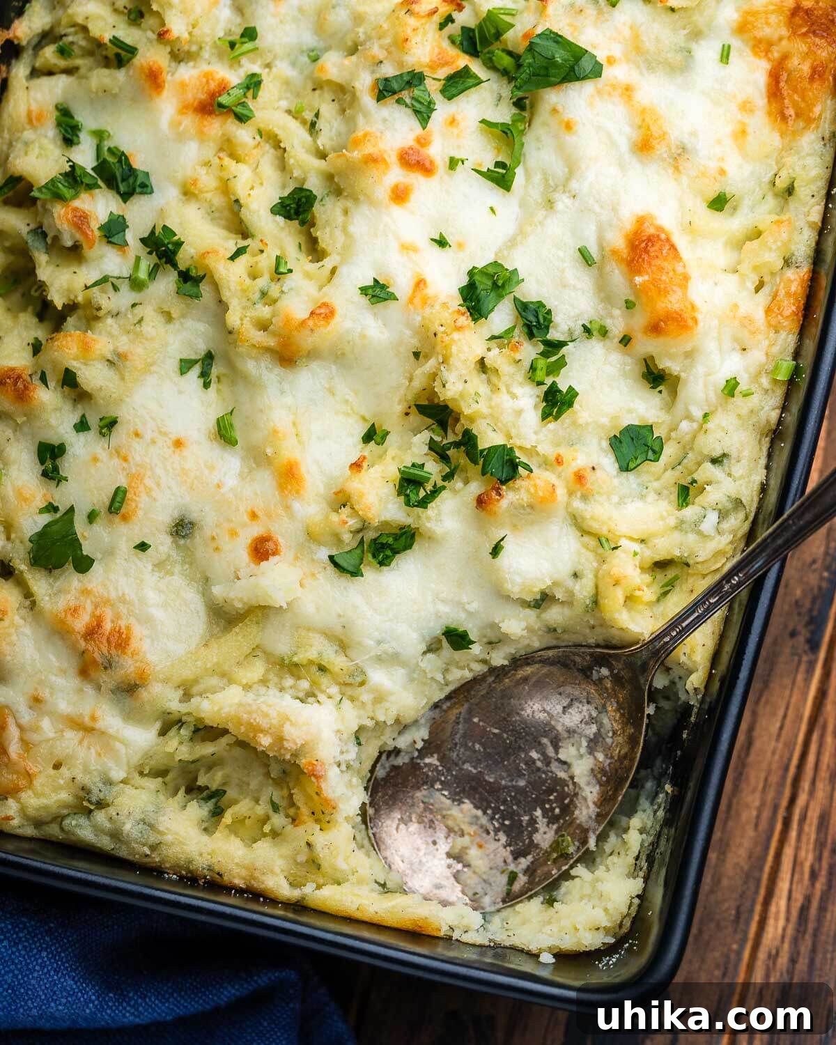 Baked Parmesan Mashed Potatoes 2 A black baking dish showcasing golden-brown Italian baked mashed potatoes, ready to be served, with a spoon beside it.