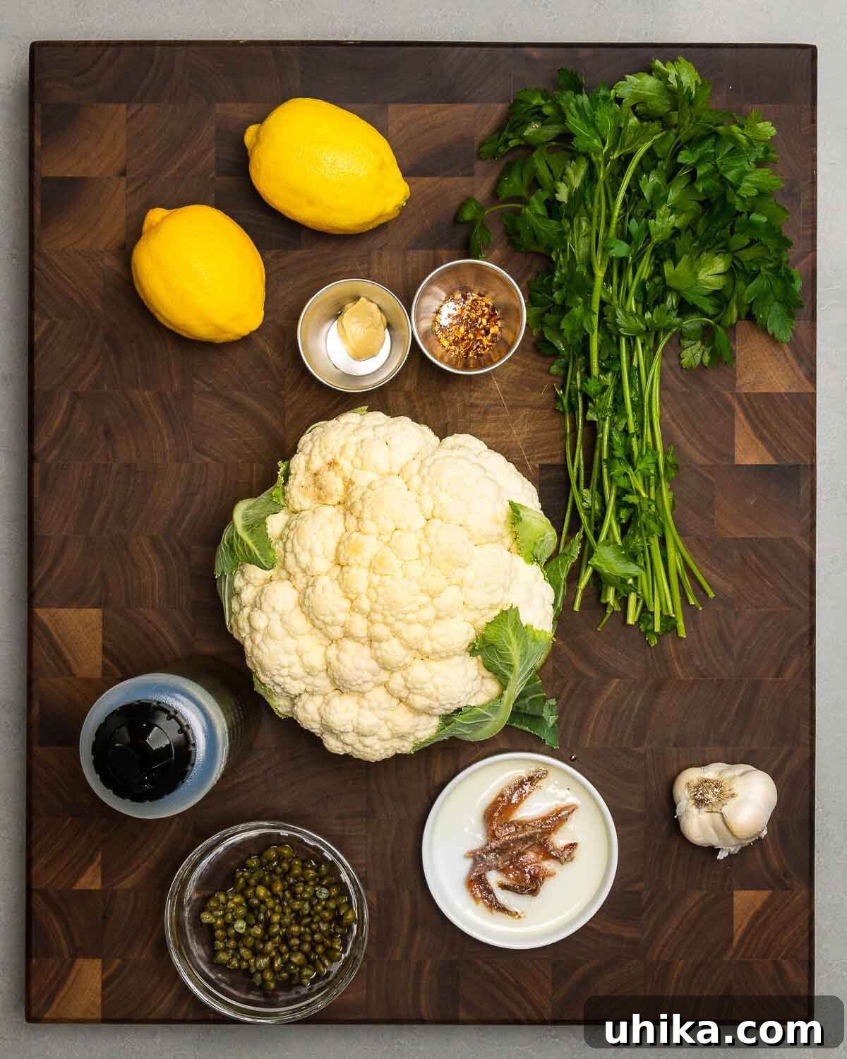Ingredients for Italian cauliflower salad laid out: fresh lemons, Dijon mustard, hot red pepper flakes, vibrant parsley, crisp cauliflower florets, golden olive oil, briny capers, savory anchovies, and aromatic garlic.