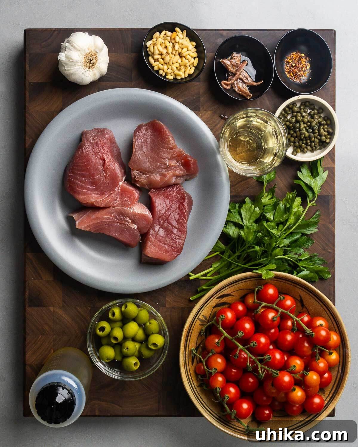 Tuna Siciliana: Olives and Capers 3 Ingredients shown: garlic, pignoli nuts, anchovies, hot red pepper, wine, capers, tuna steaks, parsley, olive oil, olives, and cherry tomatoes spread out on a wooden board.