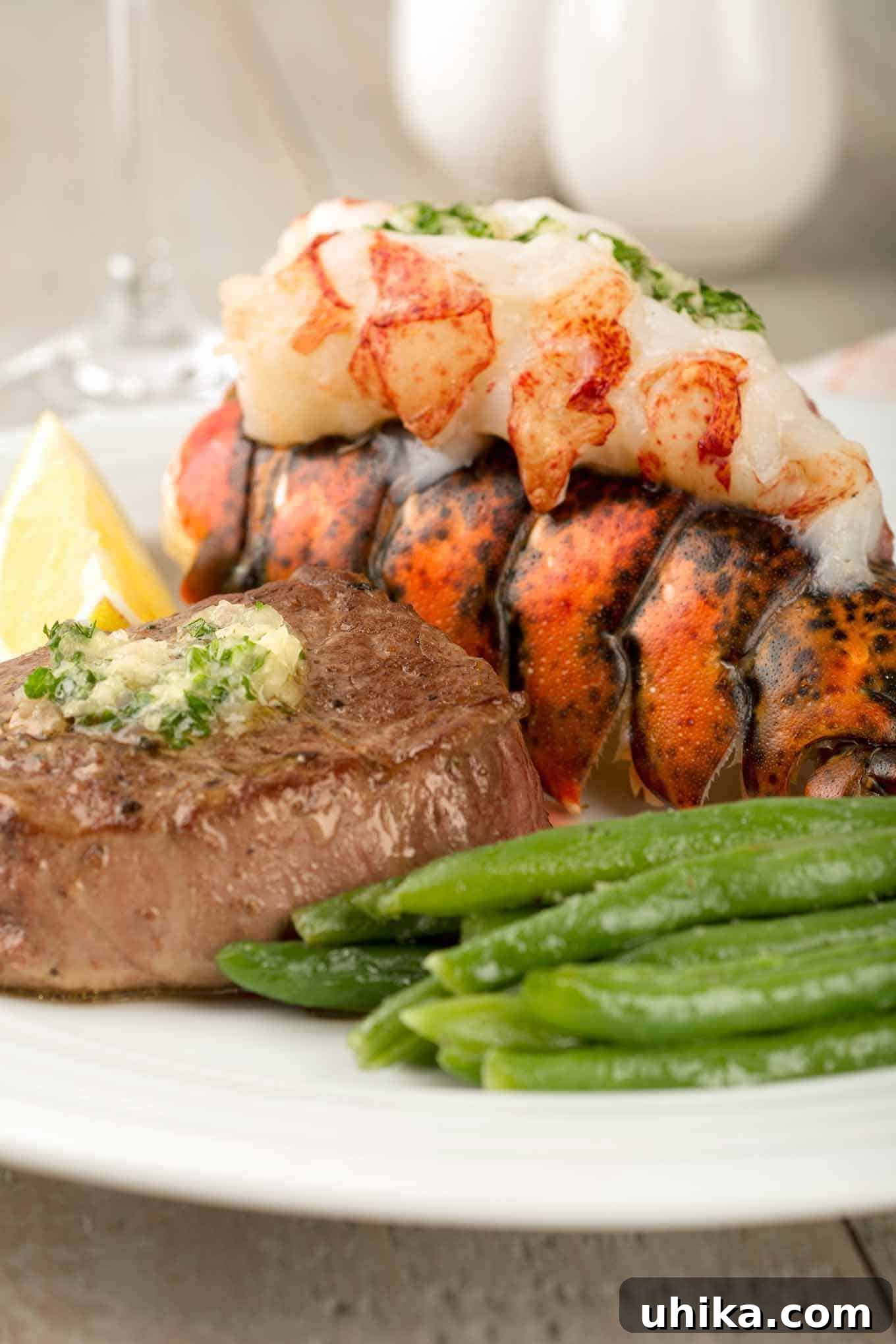 Lobster and Steak Grandeur 2 A closeup view of a pan-seared filet mignon topped with parsley butter next to a lobster tail with its juicy meat on top, and a serving of green beans. This is an easy recipe for a restaurant-quality surf and turf dinner at home.