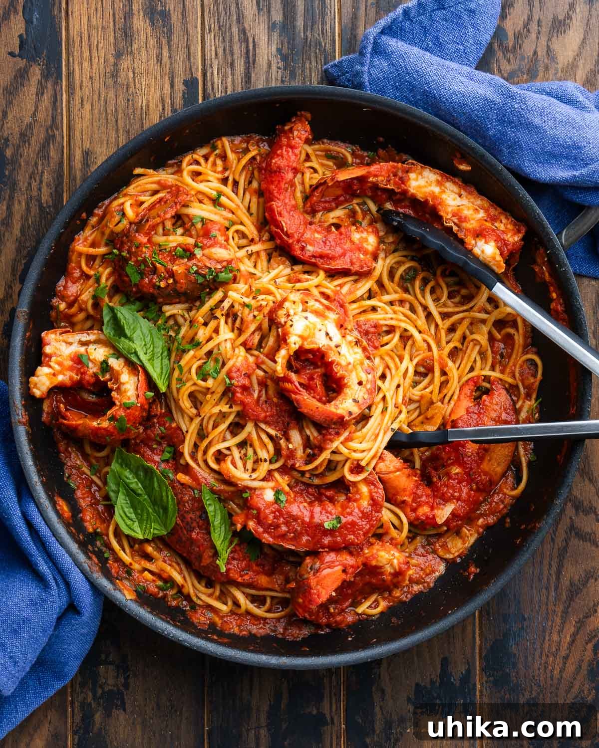 Inferno Lobster 2 Lobster fra diavolo in a black pan, garnished with fresh basil, beside two blue napkins, showcasing the vibrant colors and rich texture of the dish.