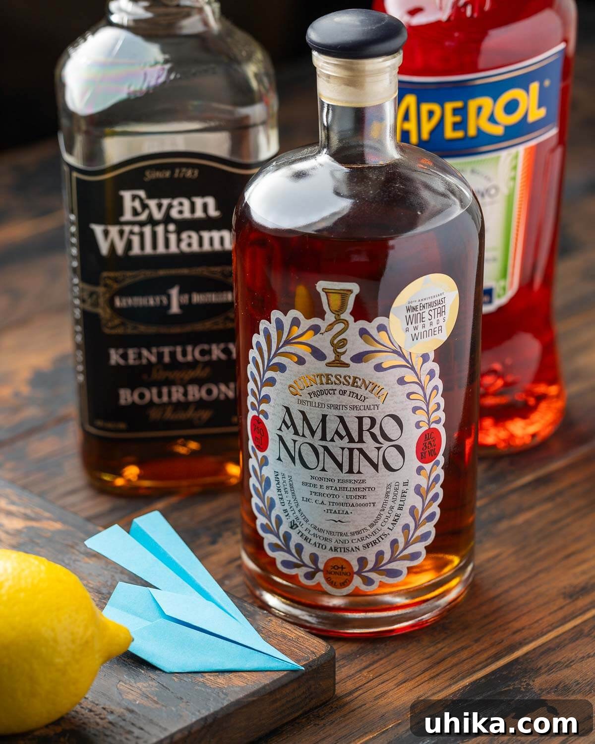 Ingredients for a Paper Plane cocktail: lemon, small blue paper planes, Amaro Nonino, bourbon, and Aperol.