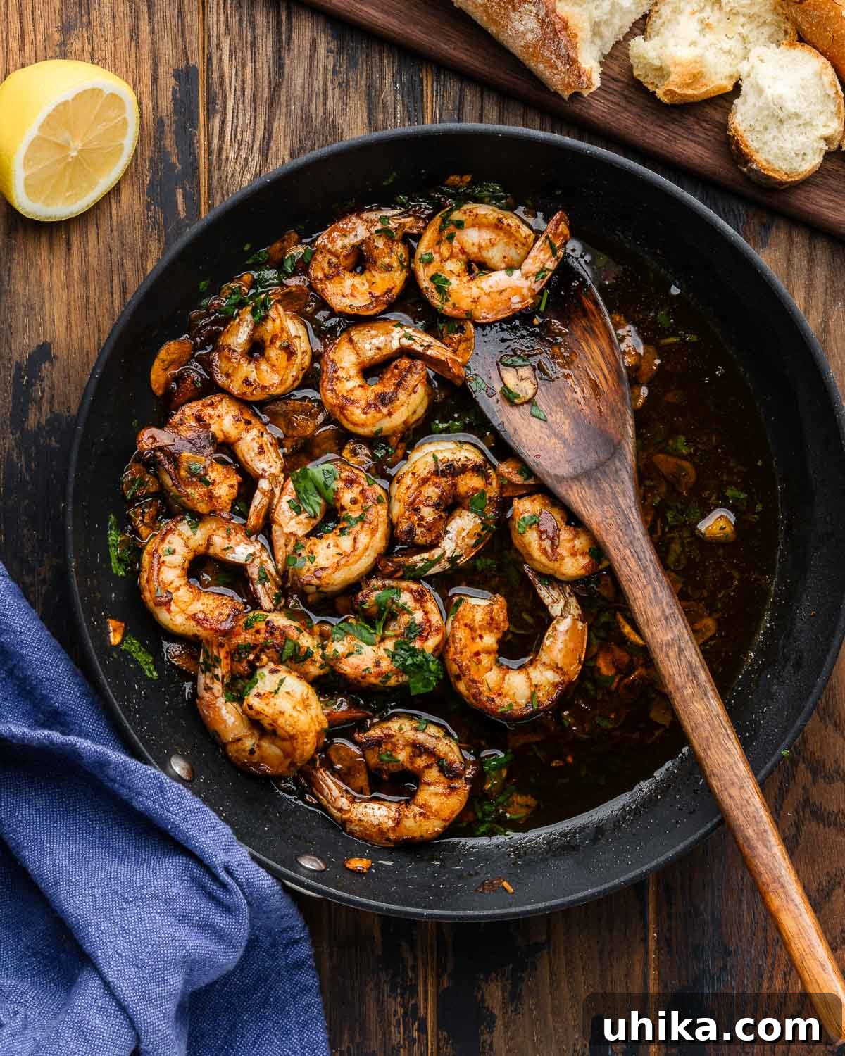 Irresistible Spanish Garlic Shrimp 2 A black pan filled with sizzling Spanish garlic shrimp, garnished with parsley, alongside lemon wedges and slices of crusty bread on a rustic wooden board.