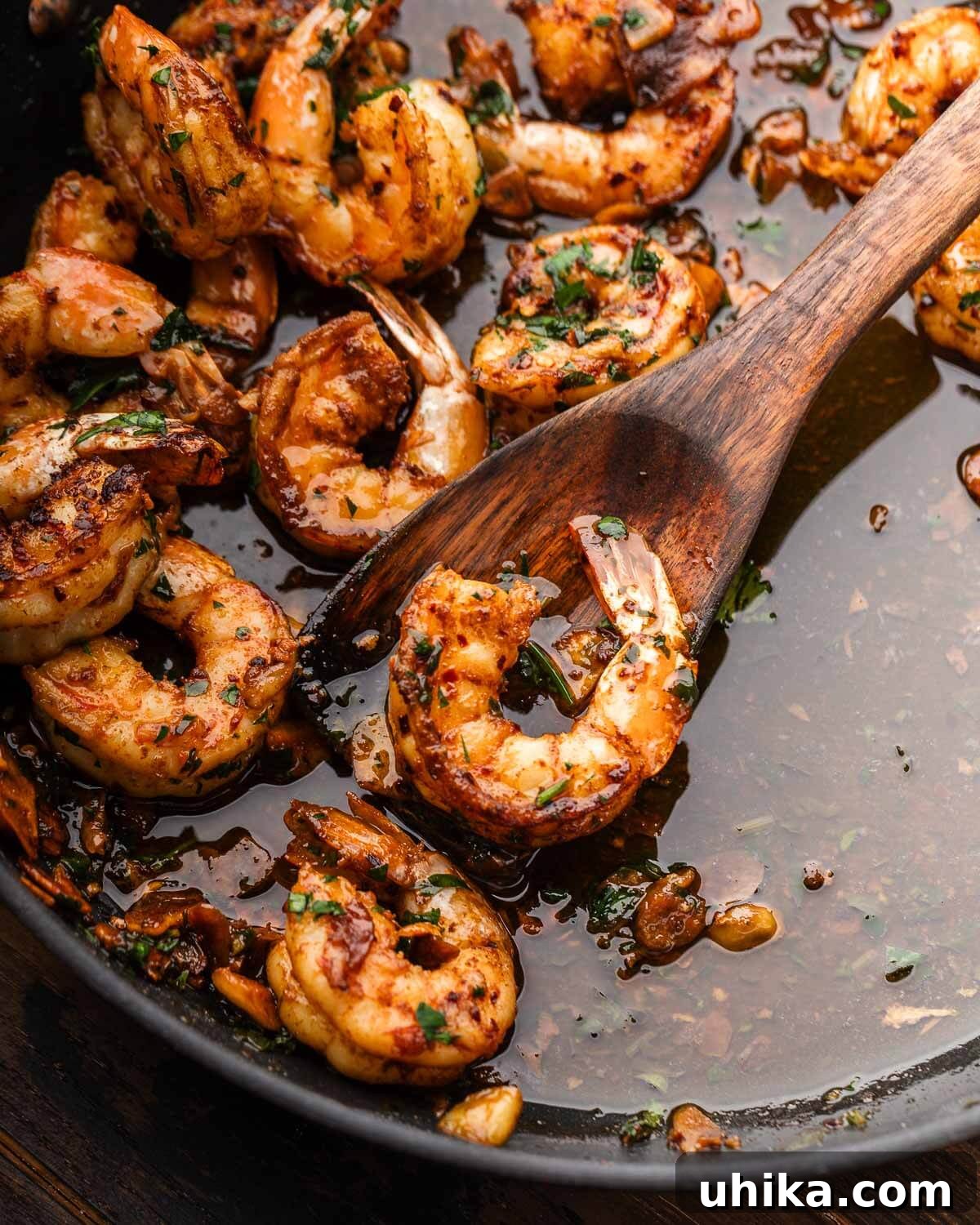 Irresistible Spanish Garlic Shrimp 5 Close-up of Gambas al Ajillo in a black pan, with a wooden spoon lifting a single shrimp, showcasing the rich garlic sauce and vibrant parsley garnish.