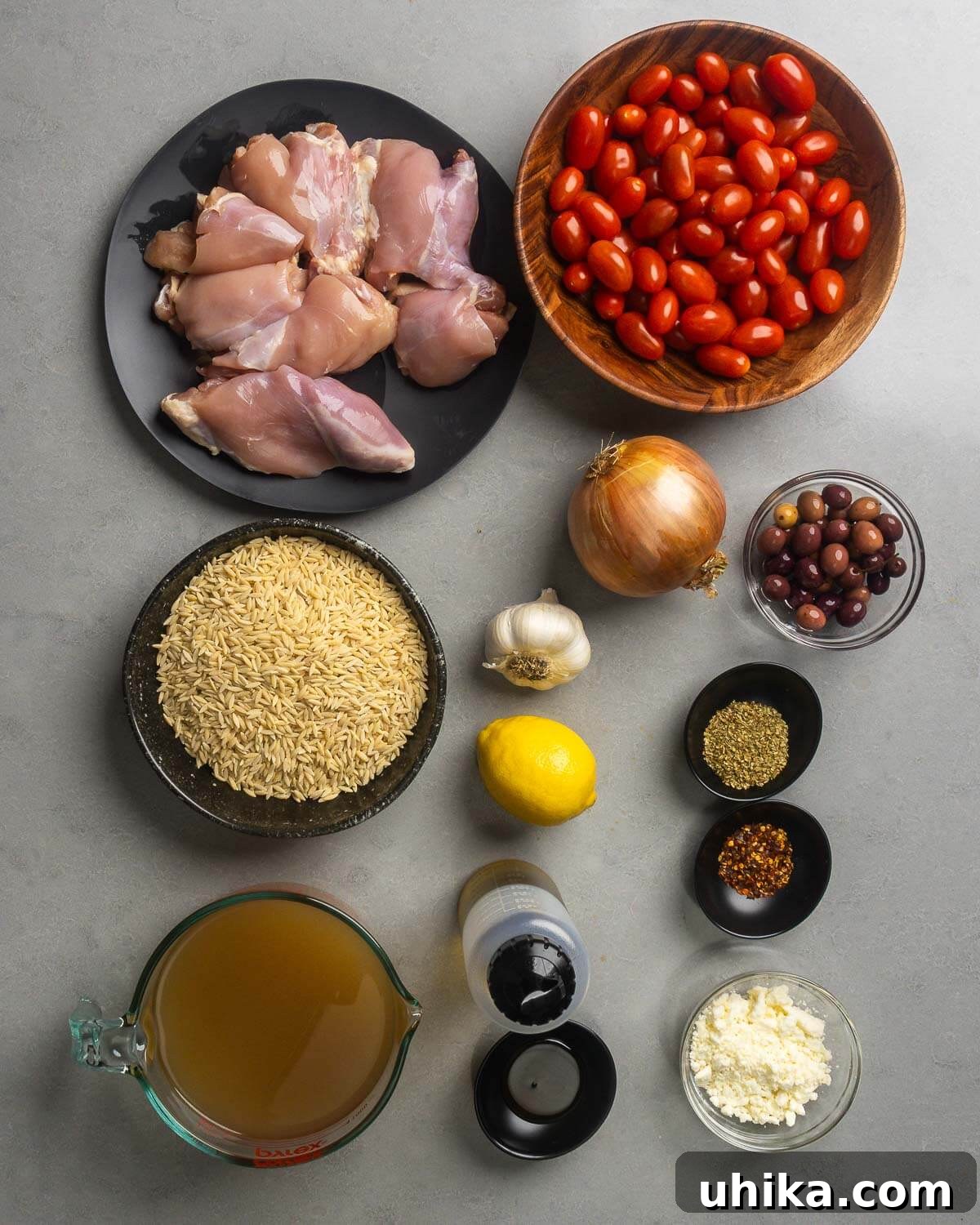 Zesty Mediterranean Chicken Orzo 3 A flat lay photograph showcasing all the fresh ingredients for Mediterranean chicken and orzo recipe: chicken thighs, cherry tomatoes, orzo pasta, garlic, onion, lemon, Gaeta olives, oregano, hot red pepper flakes, olive oil, honey, chicken stock, and feta cheese.