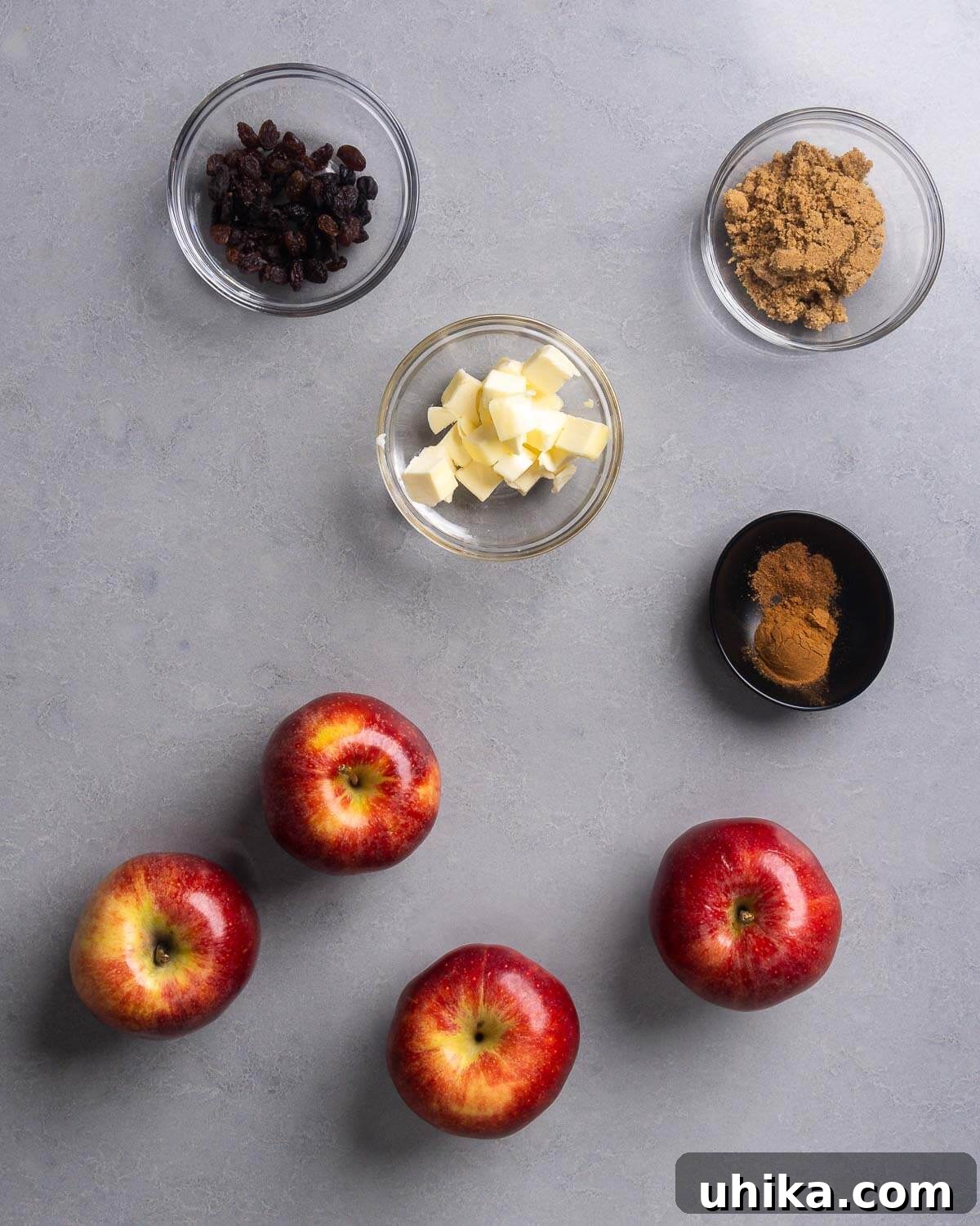 Simple Homestyle Baked Apples 3 A flat lay of ingredients for baked apples, including four apples, raisins, cubes of butter, brown sugar, cinnamon, and nutmeg.