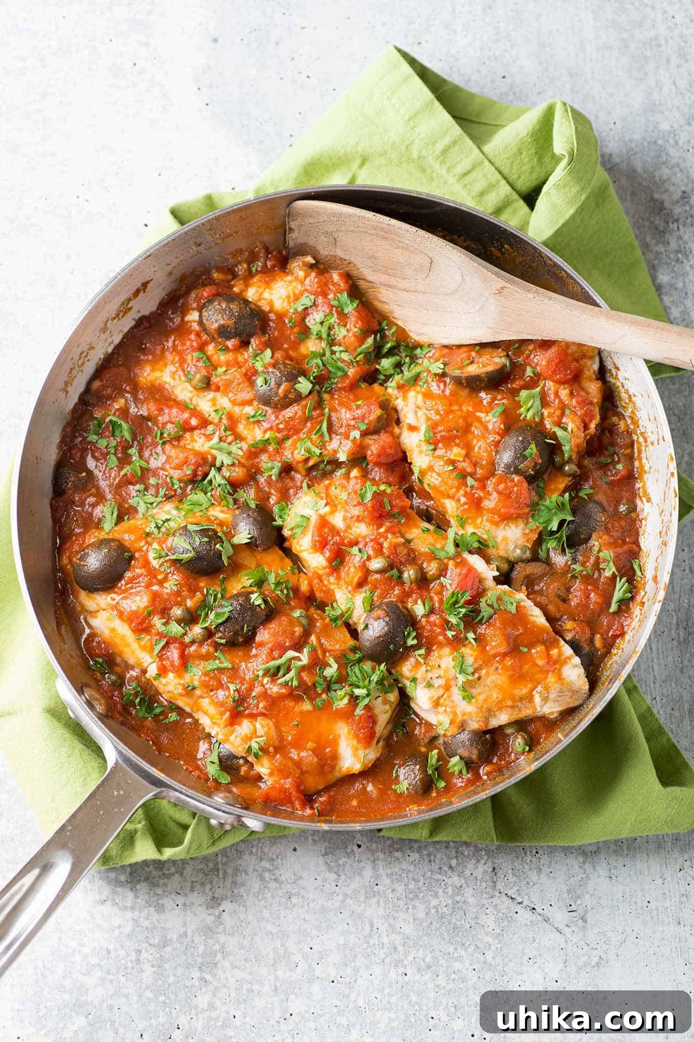 Robust Puttanesca Fish 2 An overhead view of a frying pan filled with white fish fillets in puttanesca-style tomato sauce with kalamata olives, garnished with parsley.