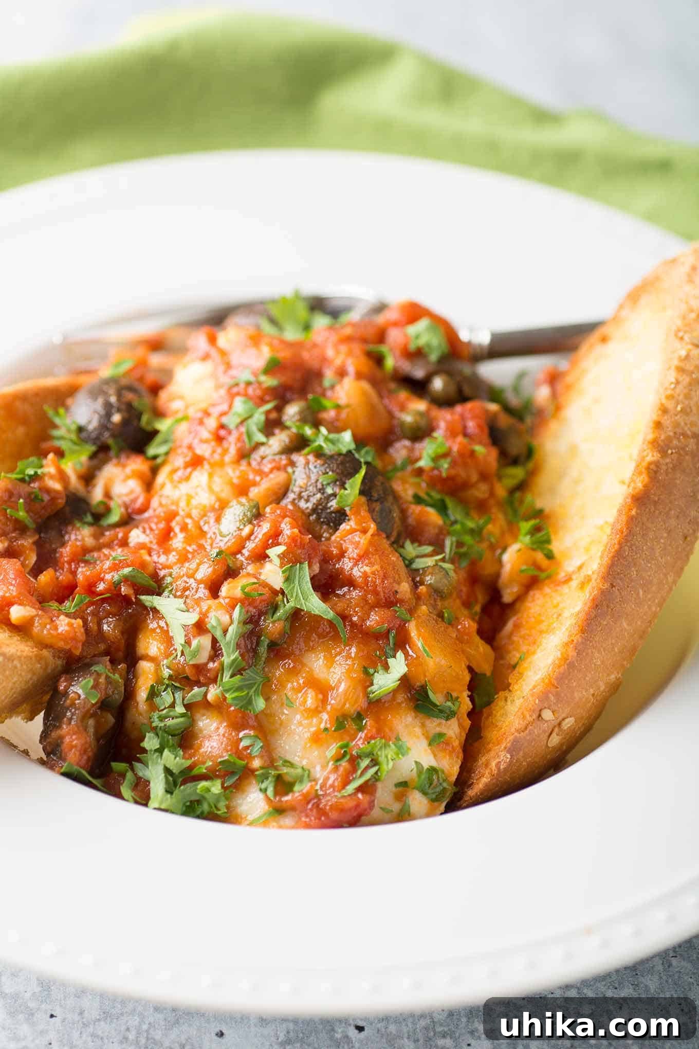 Robust Puttanesca Fish 3 A shallow bowl with a fish fillet topped with puttanesca tomato sauce made with garlic, onion, and kalamata olives, and two slices of garlic toast.