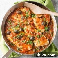 An overhead view of a frying pan filled with white fish fillets in puttanesca-style tomato sauce with kalamata olives, garnished with parsley.