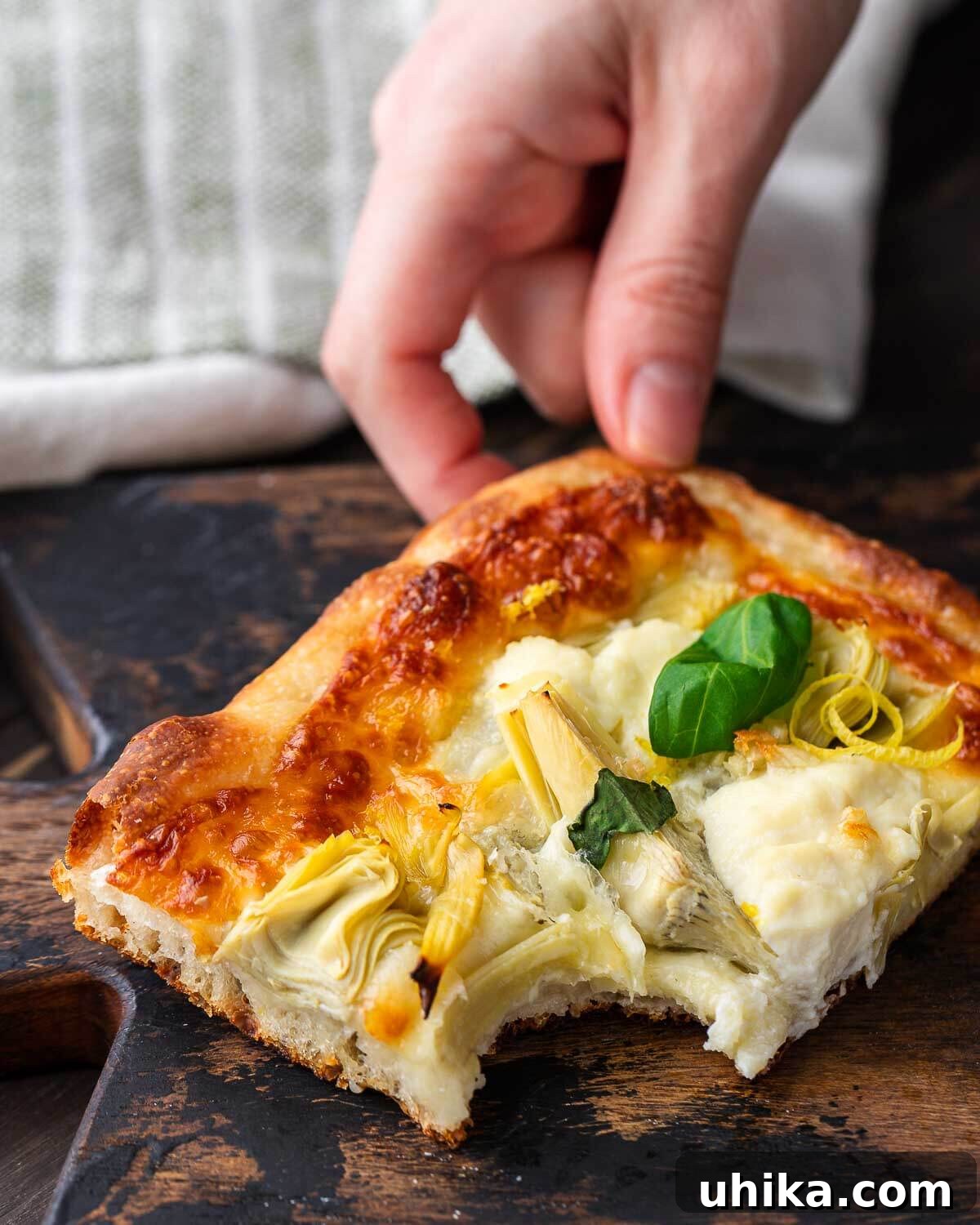 Mediterranean Artichoke Pizza 10 Hands holding a slice of artichoke pizza with a bite taken out of it, showing the cheesy interior.
