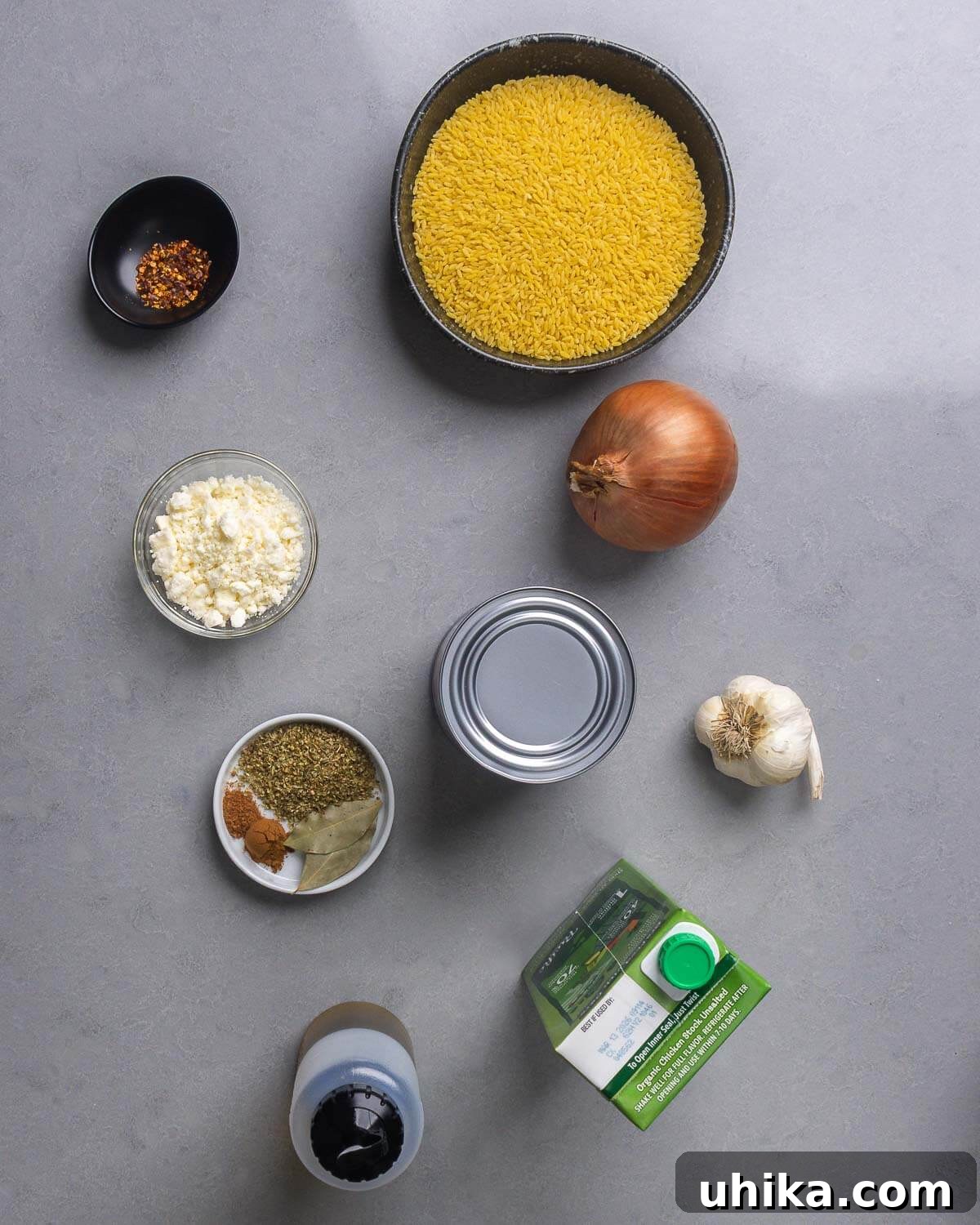 Mediterranean Orzo Bake 4 A flat lay of all ingredients required for baked orzo: hot red pepper flakes, orzo pasta, a whole onion, block feta cheese, dried oregano, ground cinnamon, ground nutmeg, bay leaves, canned plum tomatoes, fresh garlic cloves, extra virgin olive oil, and low-sodium chicken stock.