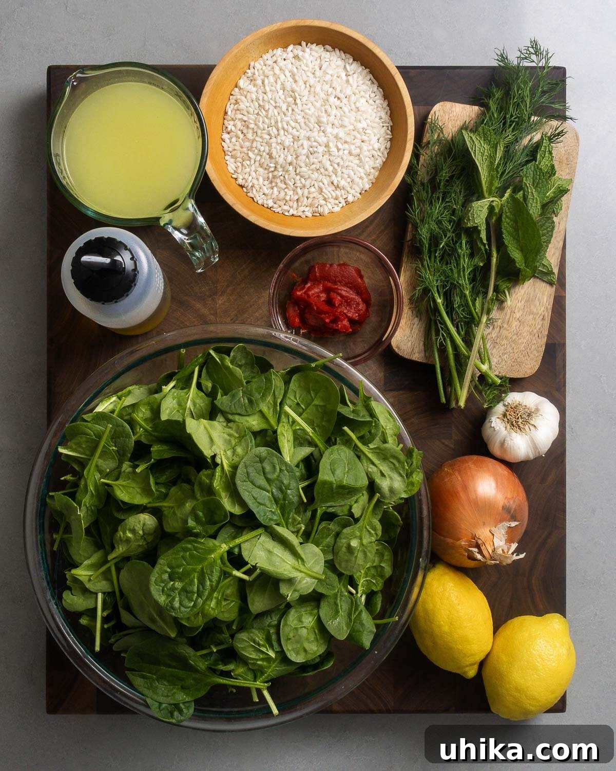 Spanakorizo: Effortless One-Pot Greek Rice 4 Ingredients shown: chicken stock, olive oil, rice, tomato paste, dill, mint, spinach, garlic, onion, and lemons, arranged on a rustic wooden board.