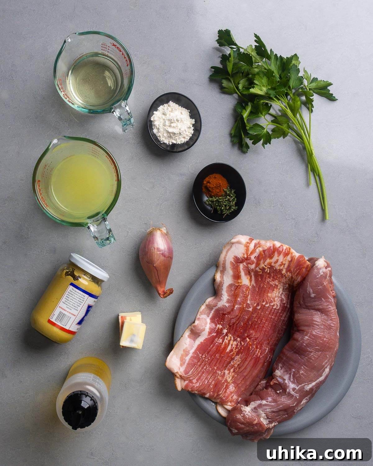 A vibrant flat lay showcasing all the fresh ingredients needed for bacon-wrapped pork tenderloin with creamy mustard sauce: wine, flour, parsley, chicken stock, paprika, thyme, shallot, Dijon mustard, butter, olive oil, bacon, and pork tenderloin.