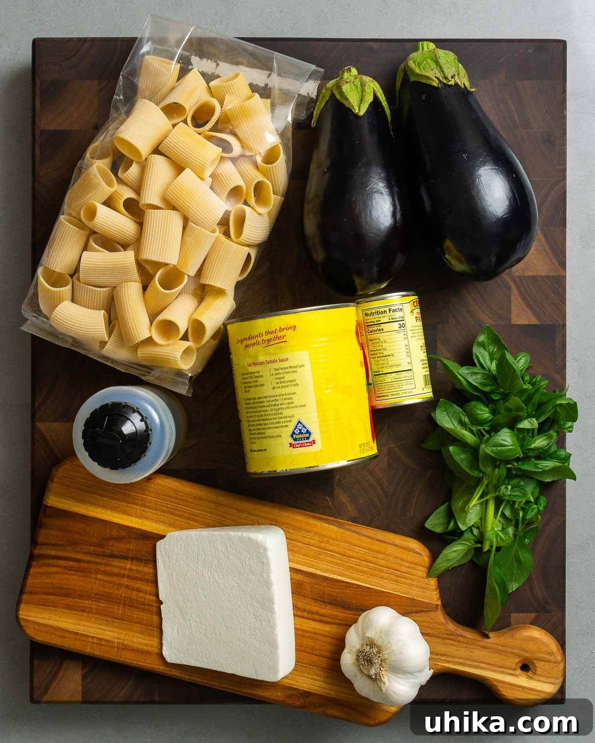 Sicilian Masterpiece Eggplant Pasta 3 A flat lay photograph showcasing all the fresh ingredients needed for Pasta alla Norma: pasta, whole eggplants, bottles of extra virgin olive oil, canned plum tomatoes, tomato paste, a bunch of fresh basil, a block of ricotta salata, and garlic cloves.