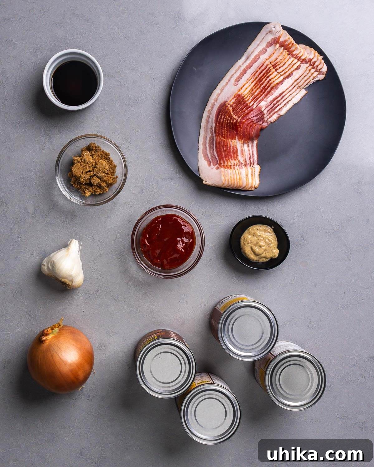 Our Signature Summer Baked Beans 4 A flat lay of all ingredients including Worcestershire sauce, brown sugar, strips of bacon, ketchup, brown mustard, fresh garlic, a yellow onion, and four cans of baked beans.