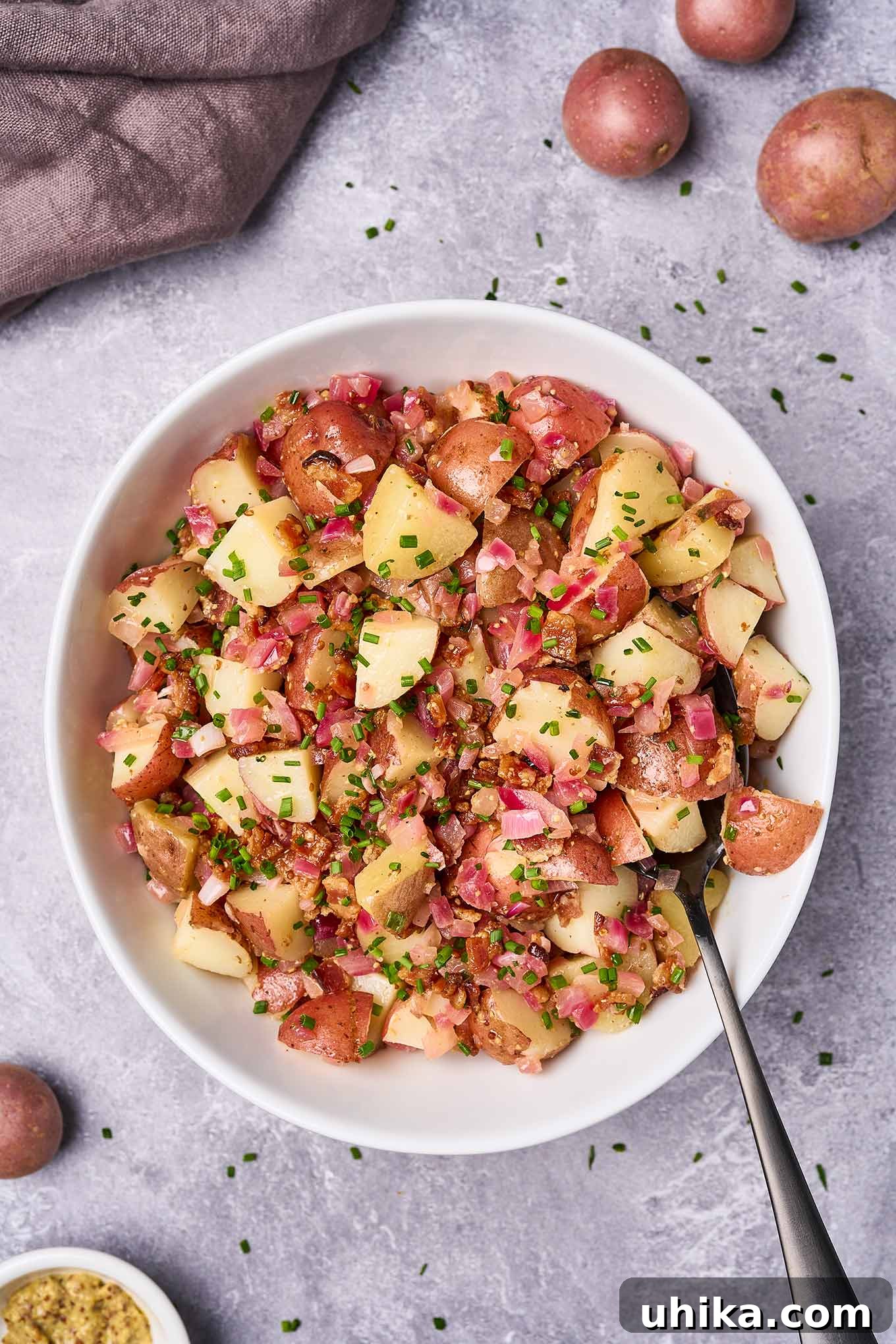 Hot Bacon Potato Salad 2 A serving bowl filled with warm potato salad with bacon dressing, serving spoon to the side, surrounded by fresh ingredients and scattered chopped chives.