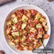 A serving bowl filled with warm potato salad with bacon dressing, serving spoon to the side, surrounded by fresh ingredients and scattered chopped chives.