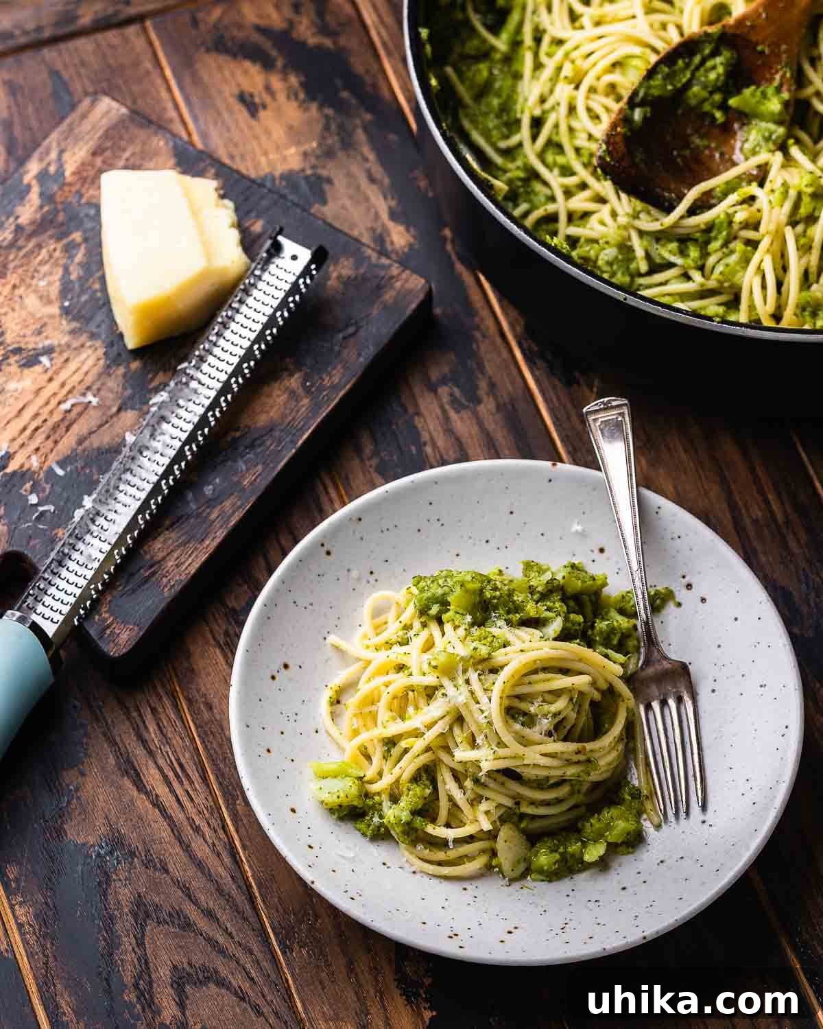 Creamy Broccoli Pasta 7 A beautifully set table featuring a white plate of spaghetti with broccoli, a large pan containing more pasta, and a wooden board with a block of Pecorino Romano cheese.