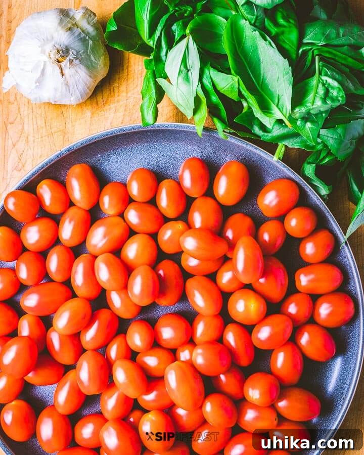 A cutting board filled with fresh ingredients: a large bowl of cherry tomatoes, a bunch of green basil leaves, and whole garlic cloves.