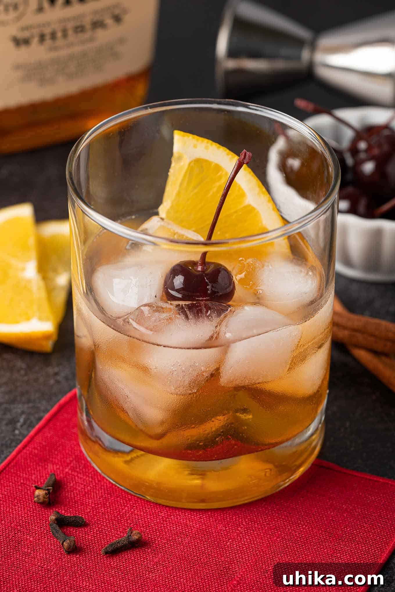 Fireside Old Fashioned 2 Old Fashioned cocktail garnished with an orange slice and a cherry, bourbon bottle, orange slices and cherries in the background.