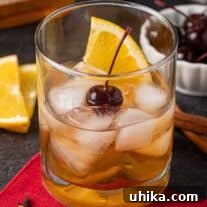 Old Fashioned cocktail garnished with an orange slice and a cherry, bourbon bottle, orange slices and cherries in the background.