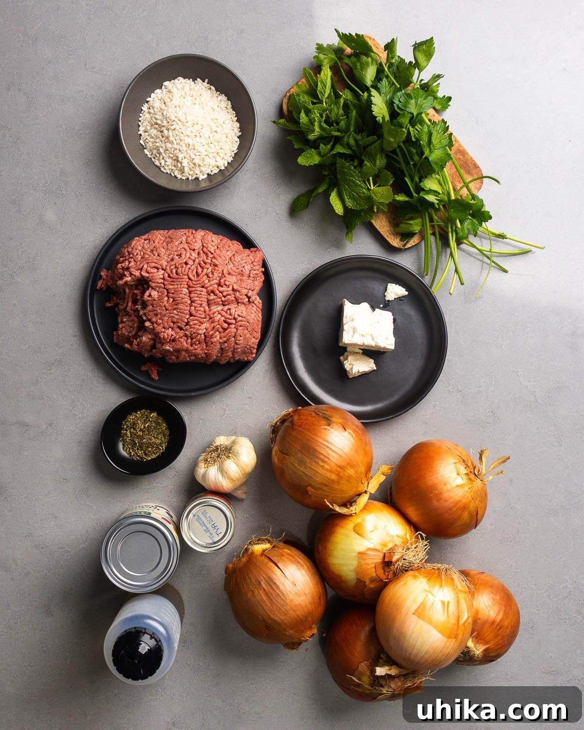 Savory Greek Onion Bake 4 A flat lay photograph displaying all the fresh and pantry ingredients for Greek stuffed onions: large yellow onions, ground chuck, arborio rice, fresh mint, fresh parsley, dried oregano, minced garlic, feta cheese, crushed plum tomatoes, tomato paste, and olive oil.