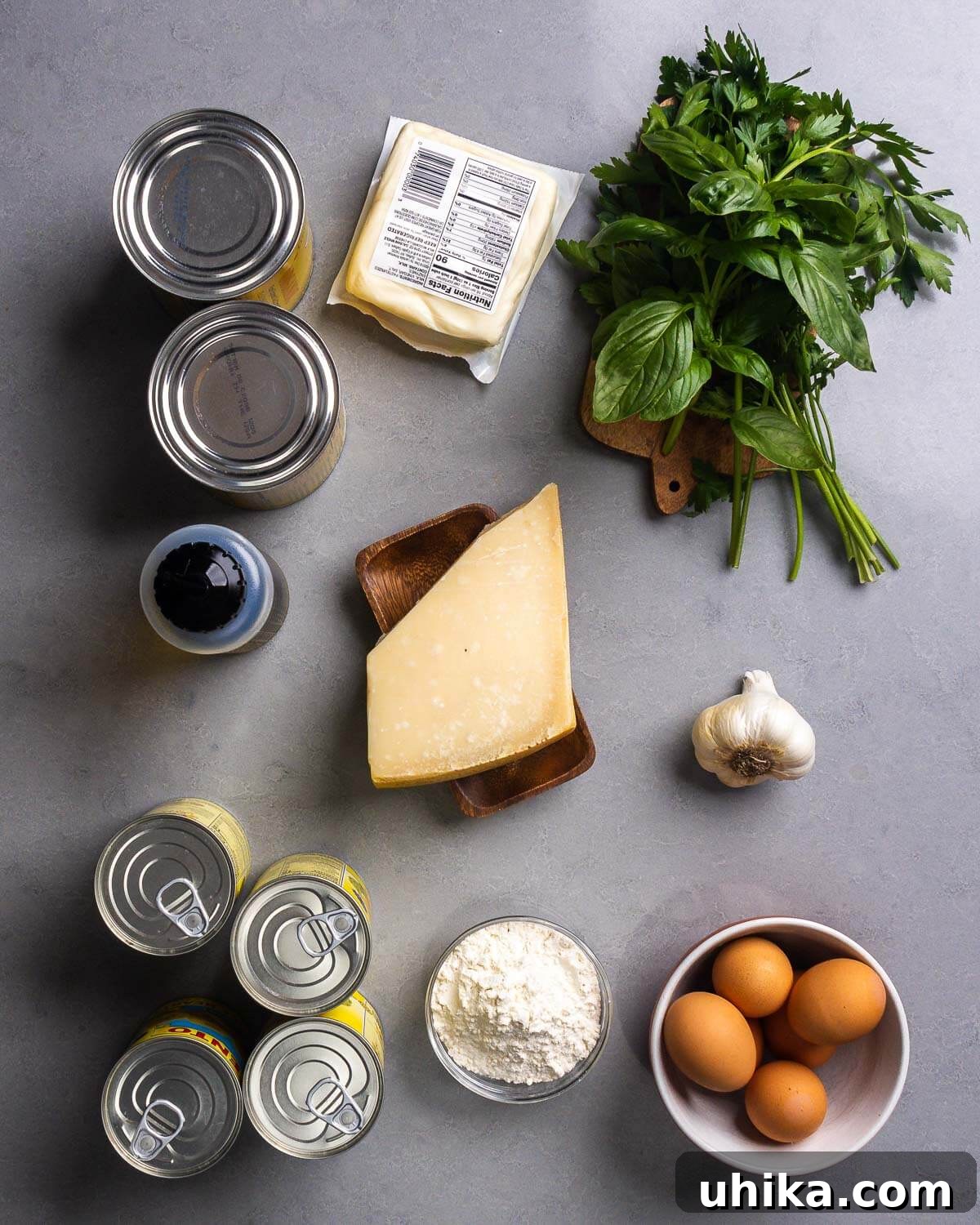Golden Artichoke Parmesan Bake 4 A flat lay photograph displaying all the ingredients for Artichoke Parmesan: canned plum tomatoes, shredded mozzarella, fresh basil, fresh parsley, olive oil, grated Parmesan cheese, garlic, canned artichoke hearts, flour, and eggs.