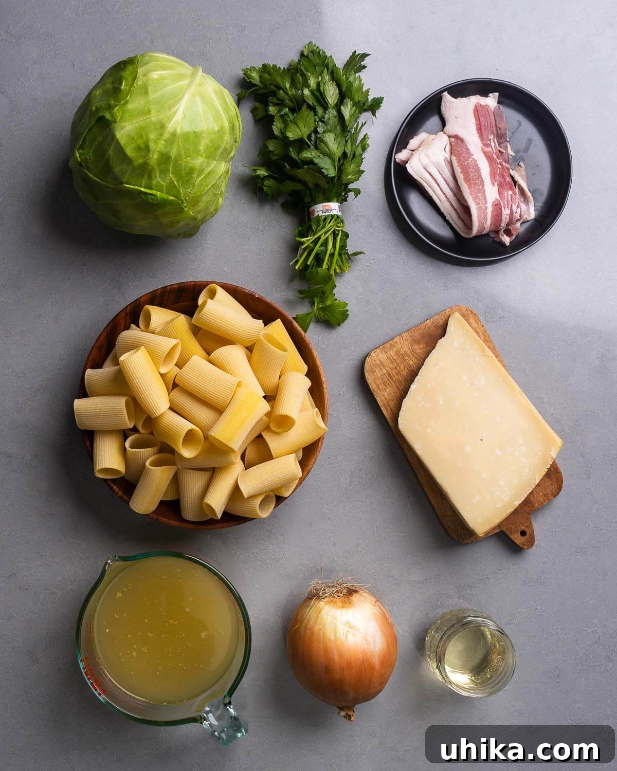 All ingredients for Cabbage Bacon Pasta displayed: cabbage, parsley, raw bacon, rigatoni pasta, a block of Parmesan cheese, chicken stock, onion, and a glass of white wine.