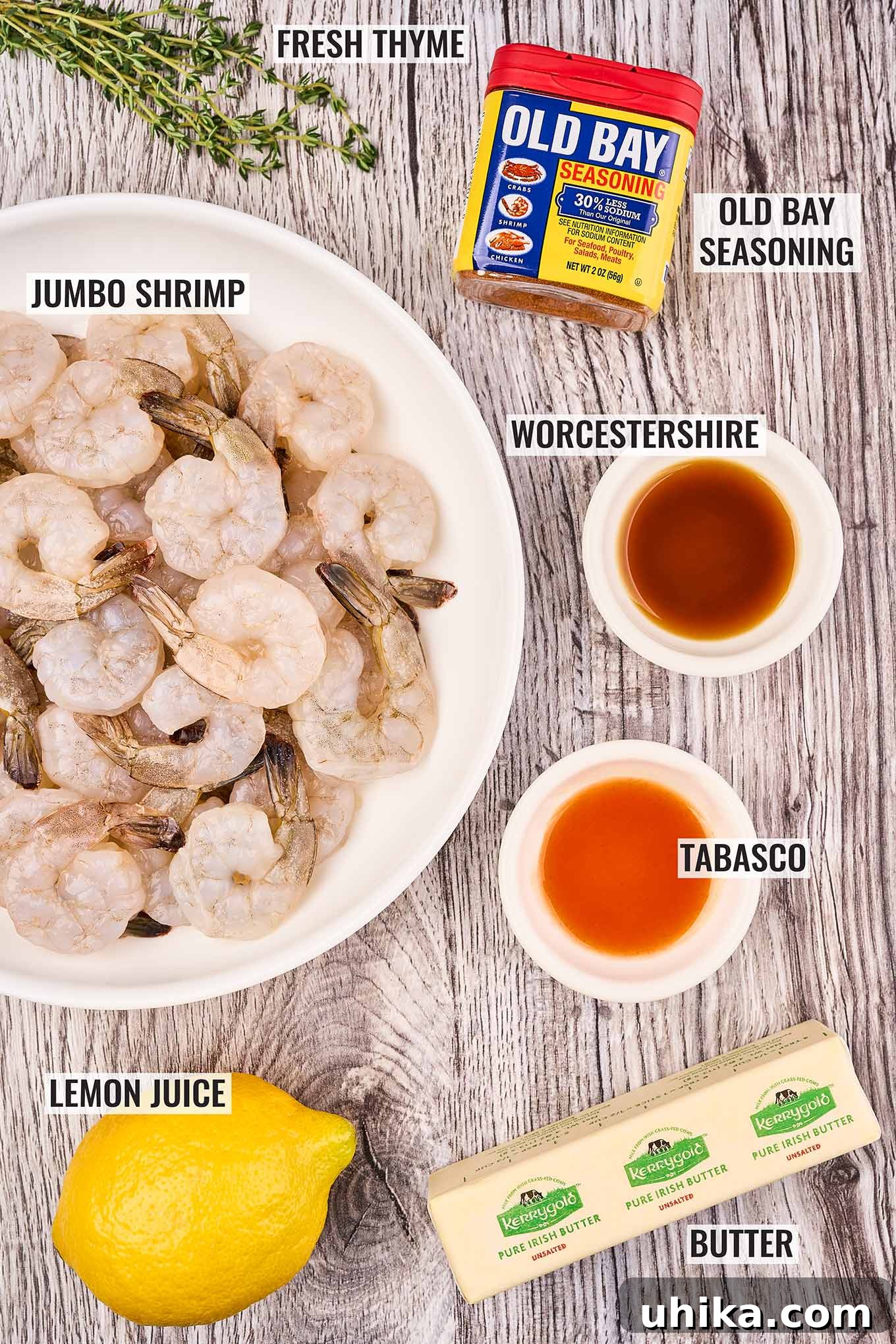 Labeled ingredients for shrimp sauté on a grey wood background. Raw shrimp, lemon, butter, Worcestershire, Tabasco sauces, Old Bay seasoning, and thyme.