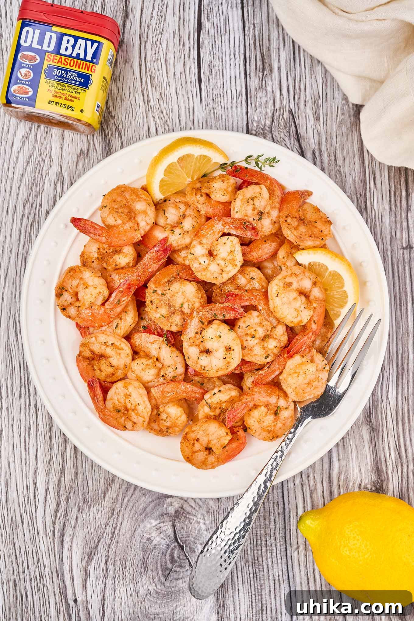 A white plate piled with Old Bay sautéed shrimp and a fork, lemon wedges, a sprig of thyme, linen napkin, and Old Bay tin in the background.