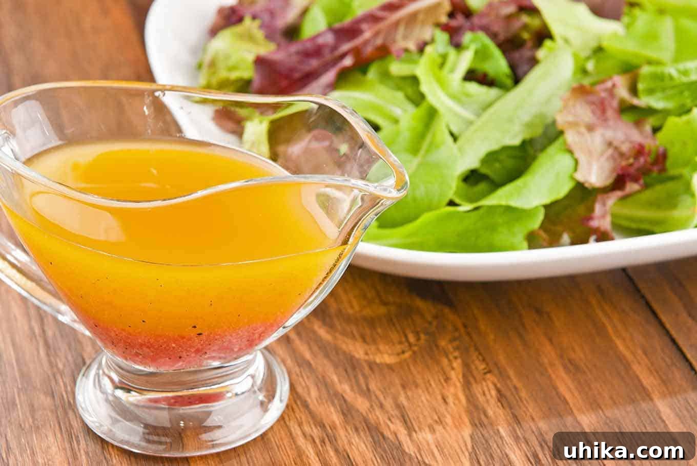 Homemade Dressing Essentials 3 A glass gravy boat filled with vinaigrette dressing in front of a plate of fresh mixed greens on a wooden table.