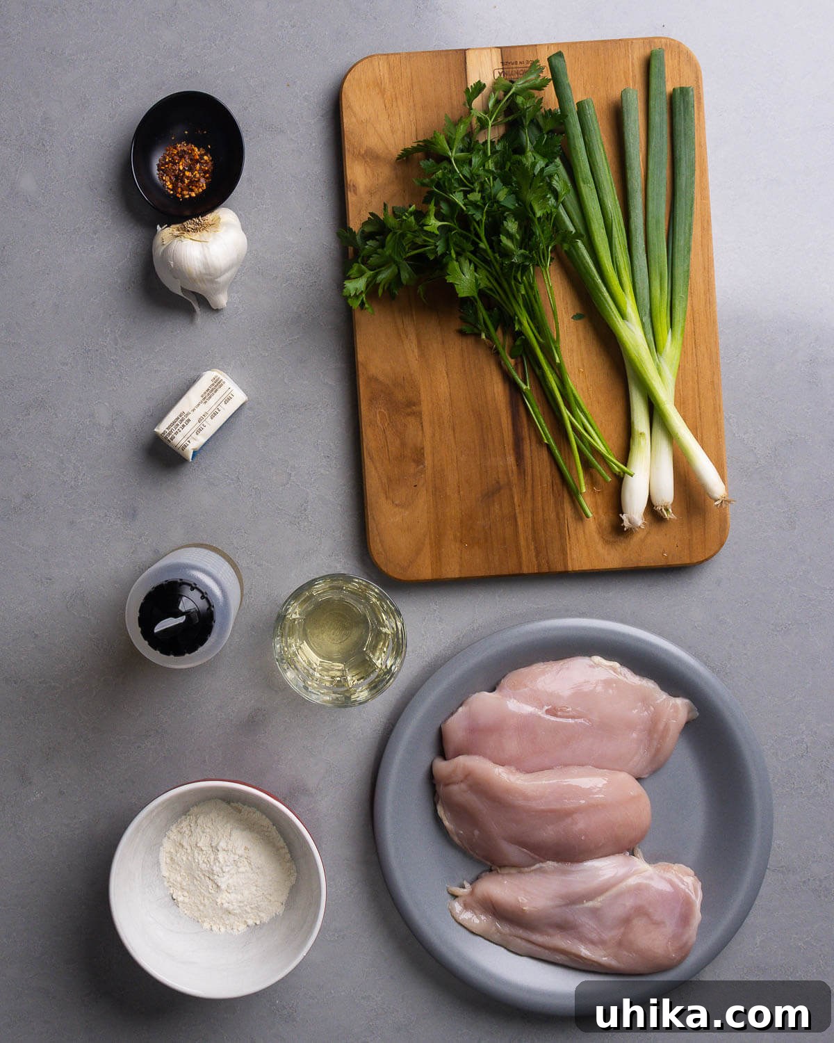 Buttery Garlic Chicken Poppers 4 A selection of fresh ingredients laid out: hot red pepper flakes, garlic cloves, fresh parsley, green onions, butter sticks, olive oil, a bottle of white wine, a bowl of flour, and raw chicken breasts.